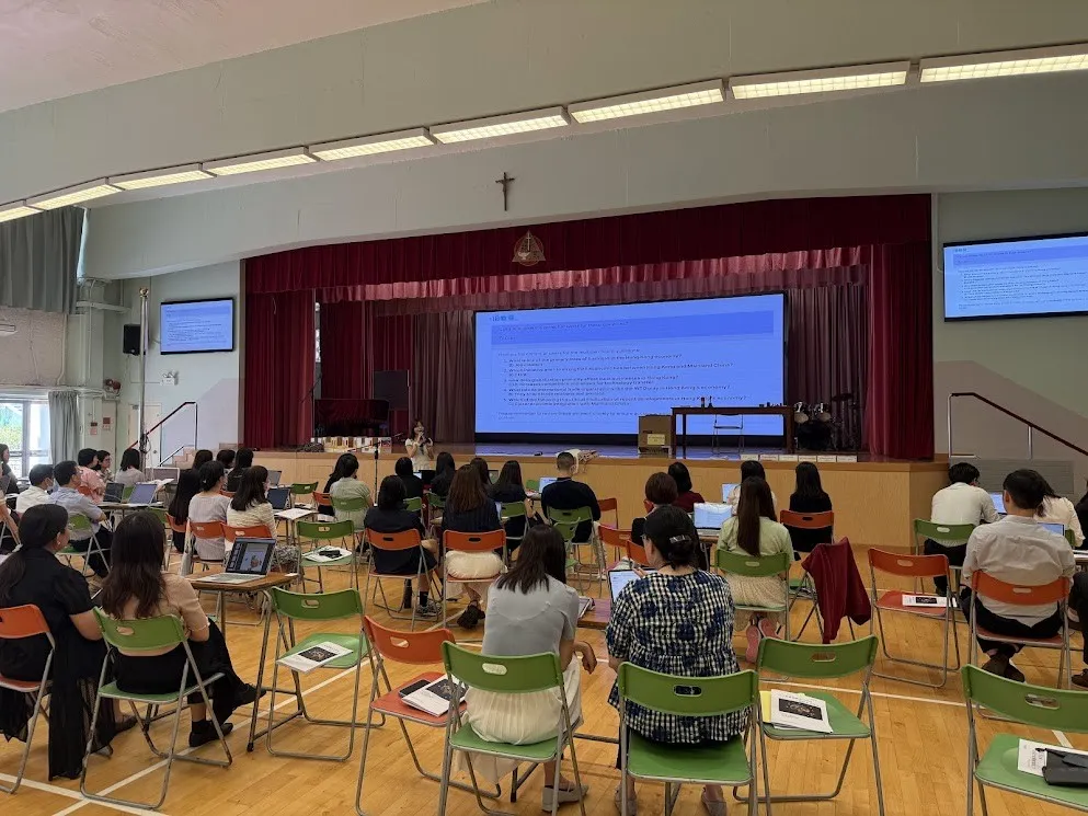 學校禮堂內，參加者席地而坐使用電腦，前方舞台設大型投影，工作坊進行中。
