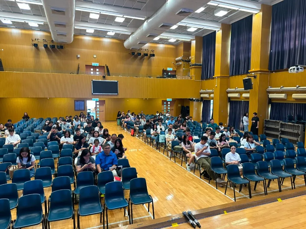 學校禮堂內舉行家長講座，大量家長坐於藍色座椅面向舞台，講台設有麥克風，展現踴躍參與的講座實況。