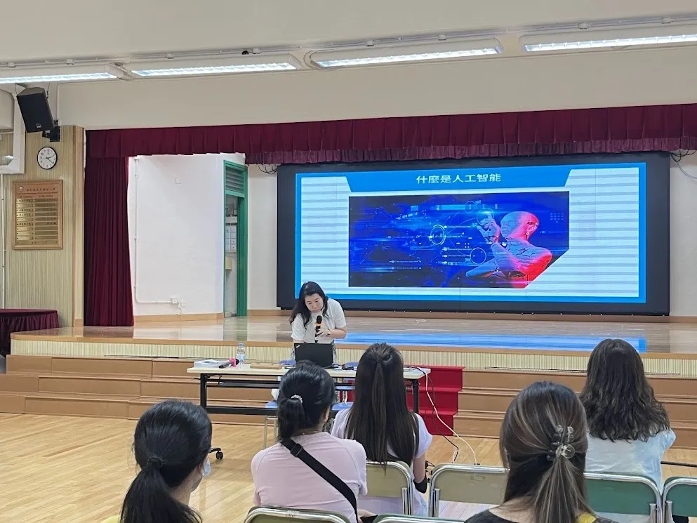 禮堂內，講者於台上分享，大螢幕顯示「什麼是人工智能」主題簡報與圖像示意。